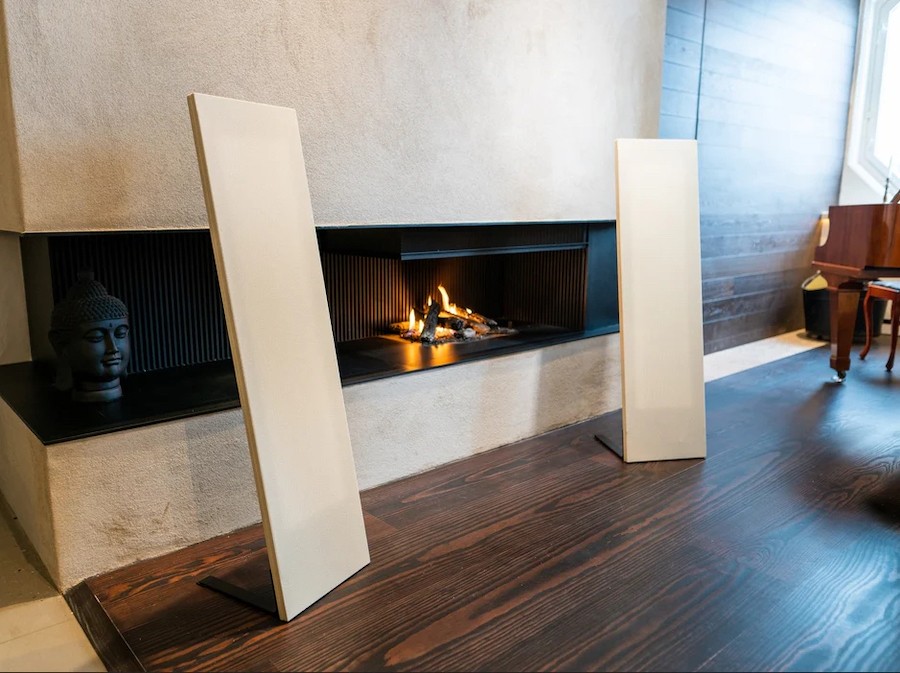 Two Magnepan speakers trimmed in beige cloth flan a modern fireplace. 